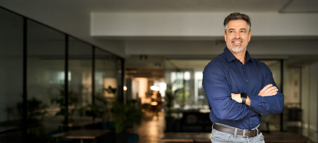 Professional headshot of a smiling man wearing a blue button-down shirt with his arms crossed in a modern office setting.