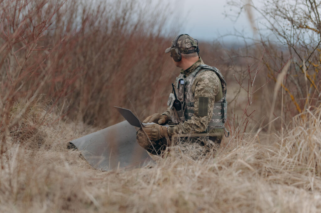 Person in camouflage using a laptop while seated in a grassy field during an outdoor operation.